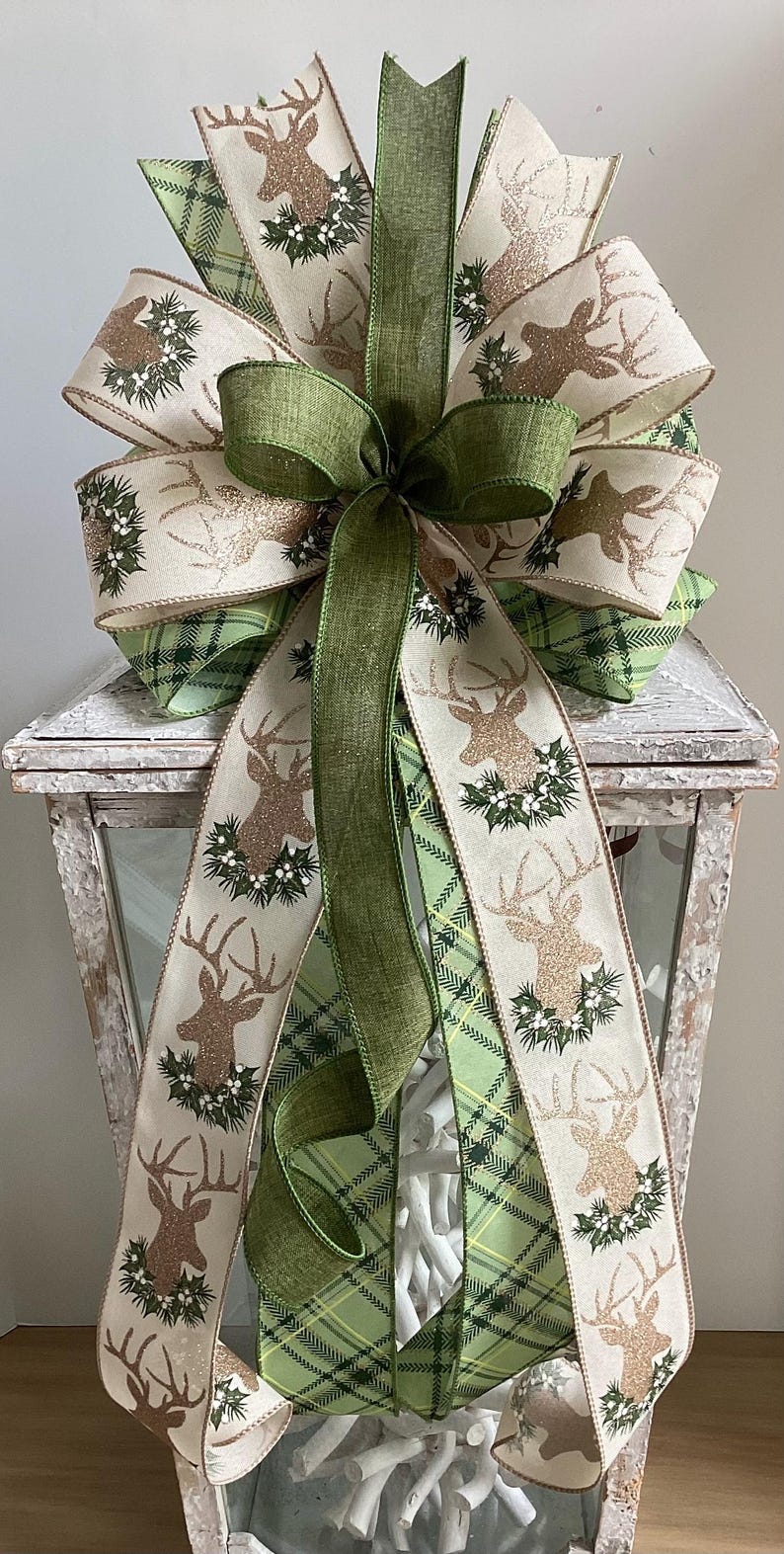 May include: A decorative bow featuring a deer and holly design. The bow has multiple layers of cream and green ribbons, with a plaid pattern and deer silhouettes. The bow is accented with a green bow and is suitable for holiday decor.
