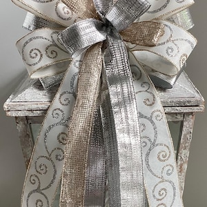 May include: A decorative bow featuring white ribbon with silver glitter swirls, silver and gold metallic ribbons. The bow is placed on top of a white, distressed lantern, creating a festive and elegant look. Perfect for holiday decor.