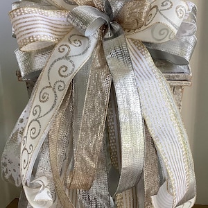May include: A large decorative bow featuring layers of white, silver, and gold ribbons. The ribbons have various patterns, including stripes, swirls, and glitter accents. The bow is likely for holiday or special occasion decor.