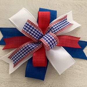 May include: A red, white, and blue bow made with gingham, satin, and burlap ribbon. The bow is tied in a knot and has a layered look.