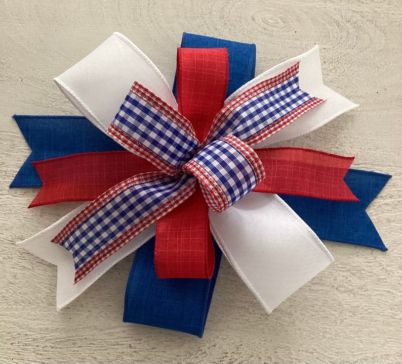 May include: A red, white, and blue bow made with gingham, satin, and burlap ribbon. The bow is tied in a knot and has a layered look.