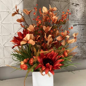 May include: A colourful floral display in a white square vase. The arrangement includes red sunflowers, orange and peach flowers, and green leaves. The background features textured white and grey panels.