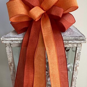 May include: A large decorative bow in shades of orange and red, with long, flowing ribbons. The bow is made of a textured fabric and is displayed on a white, distressed lantern. Perfect for fall decor.