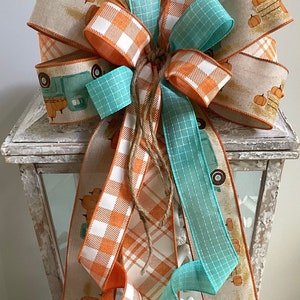 May include: A fall-themed lantern decoration with a large bow made of orange and white plaid ribbon, teal ribbon, and burlap ribbon with a vintage truck and pumpkin print.