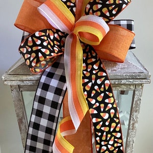 May include: A decorative Halloween bow featuring a variety of ribbons. The bow includes black and white buffalo check, orange, and ribbons printed with candy corn on a black background. The bow is attached to a white lantern.