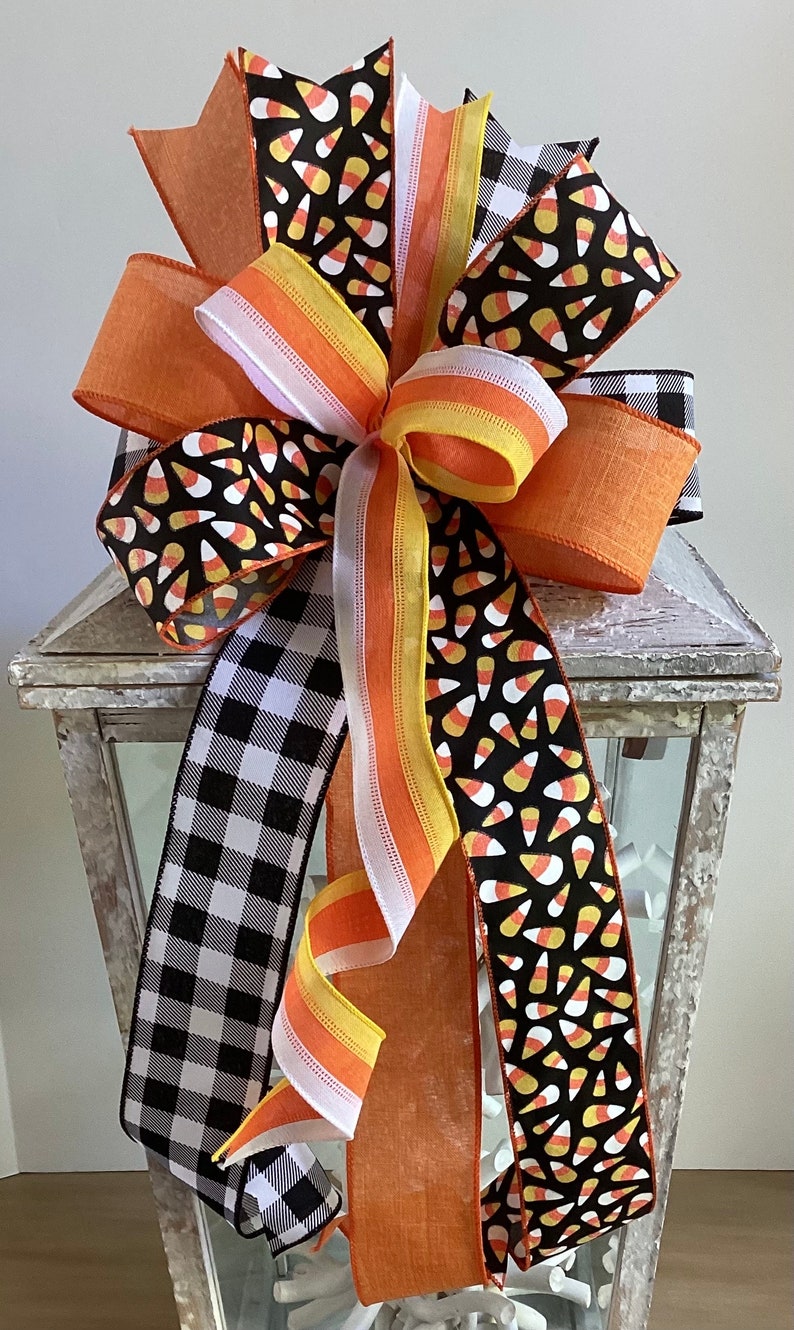 May include: A decorative Halloween bow featuring a variety of ribbons. The bow includes black and white buffalo check, orange, and ribbons printed with candy corn on a black background. The bow is attached to a white lantern.