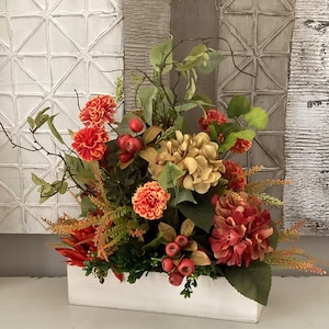 May include: A floral arrangement in a white rectangular wooden box. The arrangement features orange and red flowers, green leaves, and berries. The background includes textured panels in shades of white and gray.