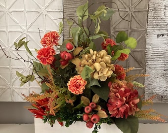 May include: A floral arrangement in a white rectangular wooden box. The arrangement features orange and red flowers, green leaves, and berries. The background includes textured panels in shades of white and gray.