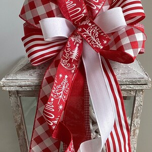 May include: A decorative Christmas bow with red and white ribbons. The bow features various patterns, including stripes, gingham, and a Christmas tree design. The bow is placed on top of a white lantern.