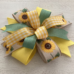 Könnte beinhalten: Dekorative Schleife mit Sonnenblumenmuster. Die Schleife hat gelbe, grüne und beige Bänder. Einige Bänder haben ein gelb-weißes Vichy-Muster. Geeignet für Kränze und Bastelarbeiten.