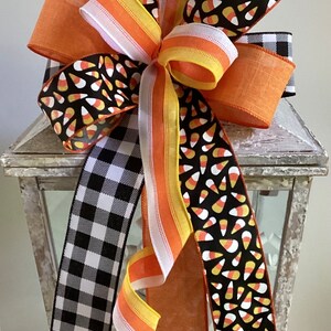 May include: A decorative Halloween bow featuring a variety of ribbons. The bow includes orange, black and white buffalo check, and candy corn patterned ribbons. The bow is a festive decoration for the fall season.