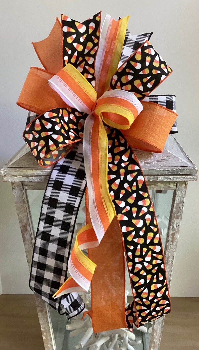 May include: A decorative Halloween bow featuring a variety of ribbons. The bow includes orange, black and white buffalo check, and candy corn patterned ribbons. The bow is a festive decoration for the fall season.