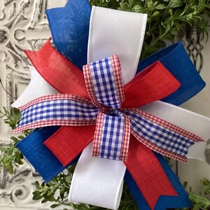 May include: A red, white, and blue bow made of ribbon with a gingham pattern. The bow is tied with a white ribbon and is perfect for a patriotic wreath or decoration.