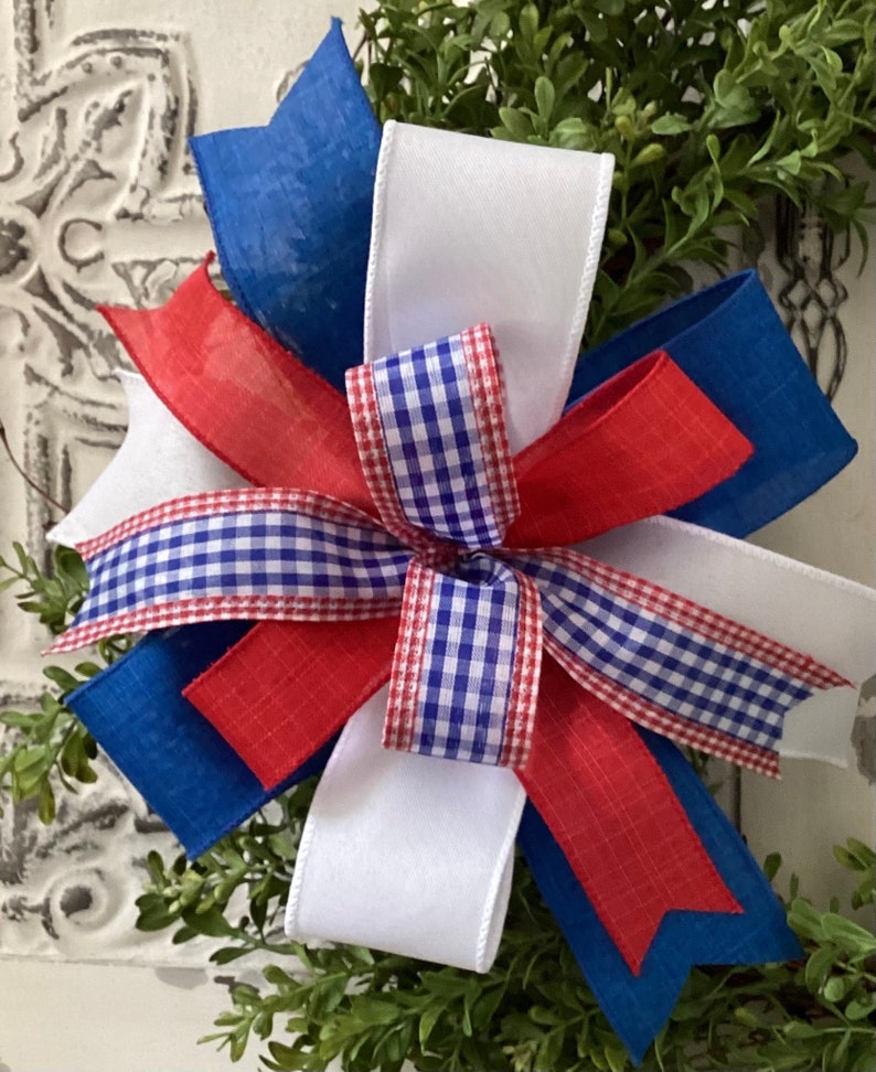 May include: A red, white, and blue bow made of ribbon with a gingham pattern. The bow is tied with a white ribbon and is perfect for a patriotic wreath or decoration.