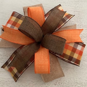 May include: A large, multi-layered bow made with orange, brown, and plaid ribbon. The bow is tied with a burlap ribbon.