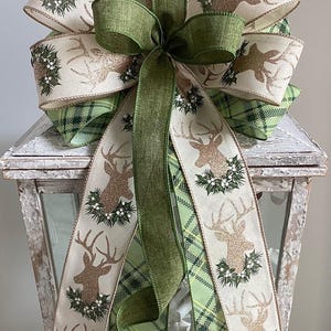 May include: A decorative bow featuring a deer design with glitter accents and a wreath. The bow has layers of cream, green plaid, and solid green ribbons. This festive bow is perfect for holiday decor.