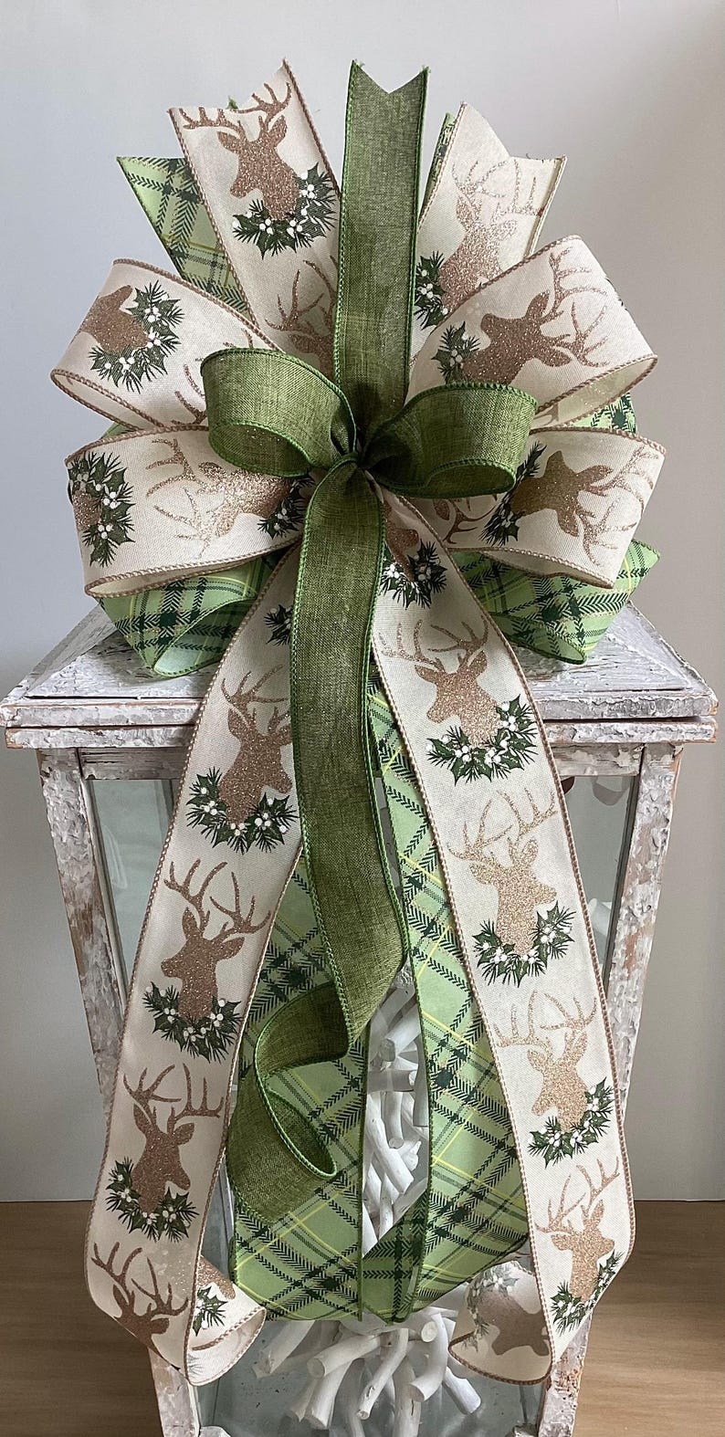 May include: A decorative bow featuring a deer design with glitter accents and a wreath. The bow has layers of cream, green plaid, and solid green ribbons. This festive bow is perfect for holiday decor.