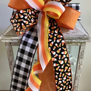May include: A decorative Halloween bow featuring orange, white, yellow, and black ribbons. The bow includes a black and white checkered ribbon, a ribbon with candy corn patterns, and solid orange ribbons. The bow is placed on top of a white lantern.