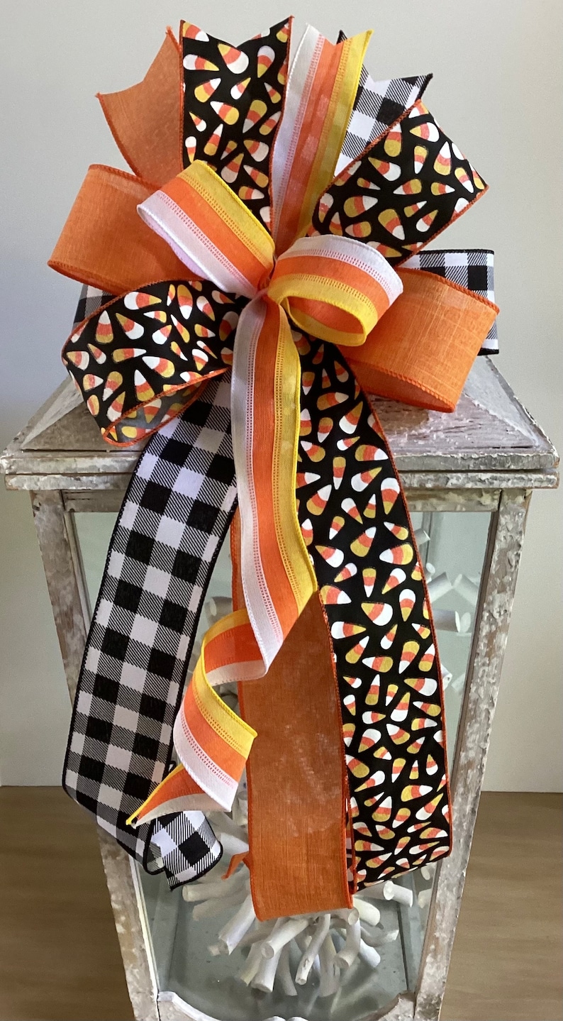 May include: A decorative Halloween bow featuring orange, white, yellow, and black ribbons. The bow includes a black and white checkered ribbon, a ribbon with candy corn patterns, and solid orange ribbons. The bow is placed on top of a white lantern.