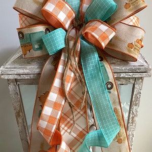May include: A fall-themed bow made with three different ribbons. The ribbons are orange and white plaid, teal blue, and a tan ribbon with a vintage truck and pumpkins design. The bow is tied with twine and is displayed on a white lantern.