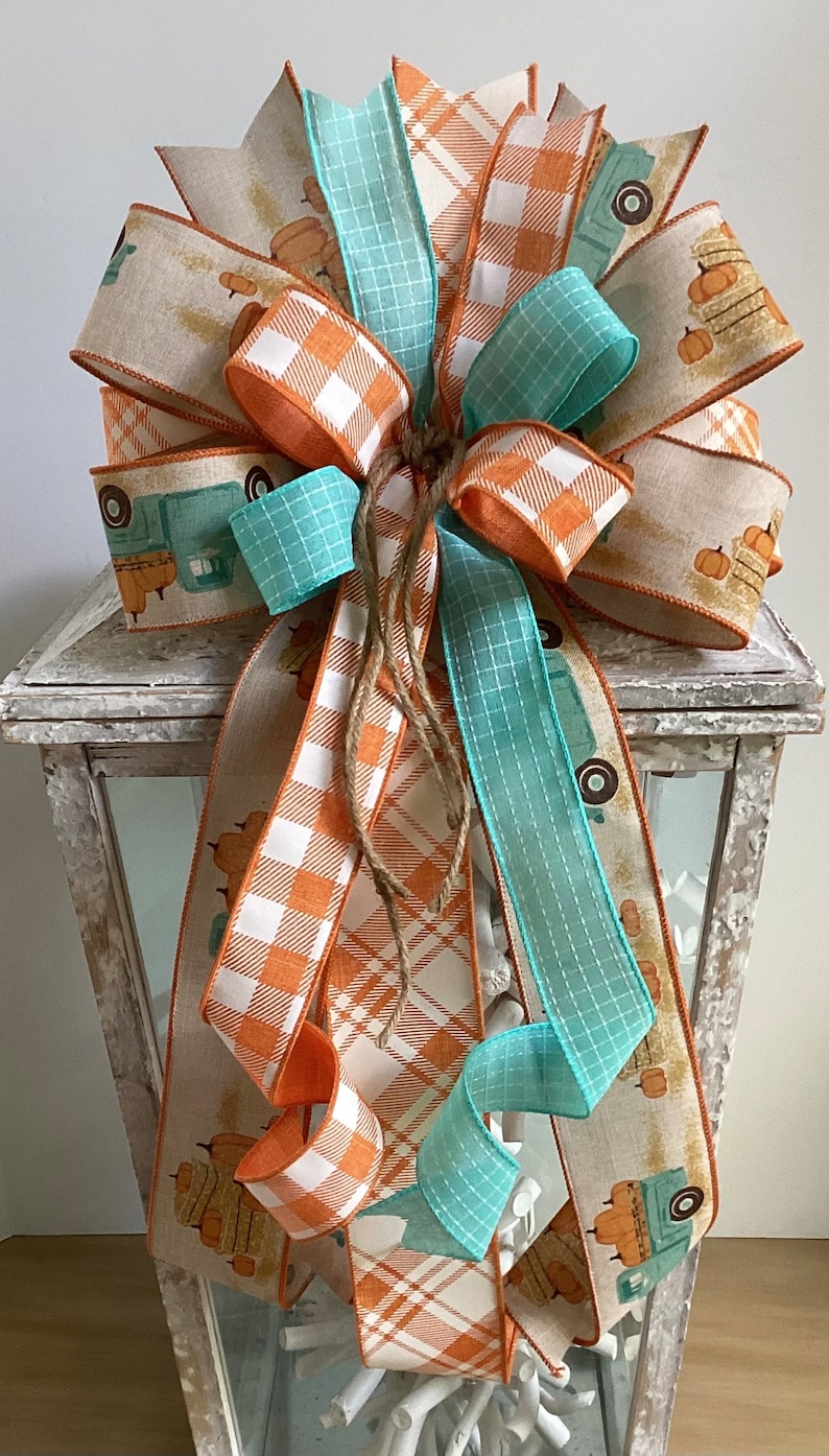 May include: A fall-themed bow made with three different ribbons. The ribbons are orange and white plaid, teal blue, and a tan ribbon with a vintage truck and pumpkins design. The bow is tied with twine and is displayed on a white lantern.