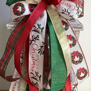 May include: A festive Christmas bow featuring multiple ribbons in red, green, gold, and white. The ribbons are adorned with Christmas wreath designs, plaid patterns, and the words "Merry Christmas". This decorative bow is perfect for holiday decor.