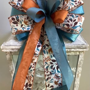 May include: A decorative bow with multiple layers of ribbon in teal, orange, and patterned fabric. The patterned ribbon features a white background with teal and brown leaves. The bow is placed on top of a white lantern.