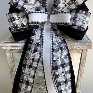 May include: A decorative bow with a black and white buffalo check design and snowflake details. The bow has long, flowing ribbons and is made from a mix of materials. Suitable for festive or winter decorations.
