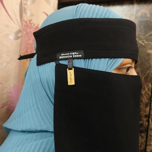 Puede incluir: Un niqab negro con una diadema a juego y un hiyab plisado azul claro. La diadema tiene el texto "BEDOON ESSM" y una etiqueta dorada con texto árabe. El niqab cubre la parte inferior de la cara.