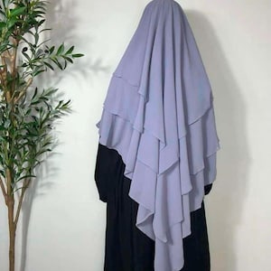 Könnte beinhalten: Ein helllilafarbenes Hijab mit mehreren Schichten, das über einer schwarzen Abaya drapiert ist.