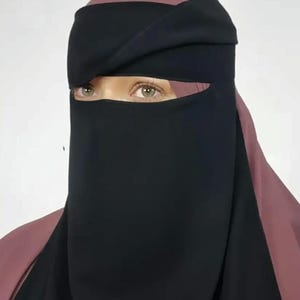 Op de afbeelding: Een zwarte niqab met bijpassende hoofdbedekking. De niqab bedekt het onderste deel van het gezicht en onthult de ogen. De hoofdbedekking is roze van kleur. Een gouden zegel met de tekst "Premium Quality" staat rechtsboven.