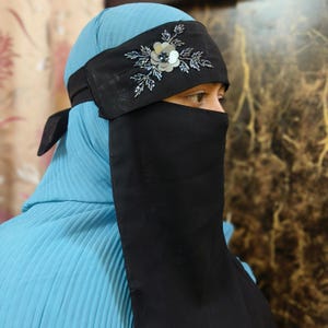 May include: A black niqab with a matching headband. The headband is adorned with a silver flower and beaded accents. The niqab is paired with a light blue pleated garment.