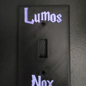 以下が含まれることがあります： 白い文字で「Lumos」と「Nox」と書かれた黒いライトスイッチカバー