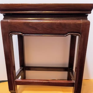 May include: A dark brown wooden plant stand with a rectangular top and a lower shelf. The stand has a simple, traditional design with a smooth finish.