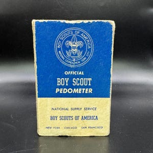May include: A vintage Boy Scout pedometer box. The top half is blue with the Boy Scouts of America emblem and text. The bottom half is tan with text that reads "National Supply Service" and "Boy Scouts of America."