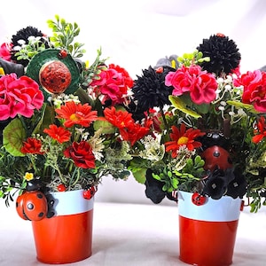 May include: Two floral arrangements in orange and white buckets. The arrangements feature red, pink, and black flowers with green foliage. Ladybug decorations adorn the buckets. The background is white.