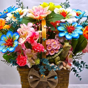 May include: A vibrant floral arrangement in a woven basket, featuring an array of colorful artificial flowers. The bouquet includes blue, orange, pink, and white blooms, accented with green foliage and a decorative turtle figurine. A burlap bow adorns the basket.