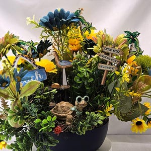 May include: A decorative floral arrangement in a dark blue pot, featuring yellow and blue flowers, greenery, and beach-themed figurines. Includes a sign that says "Beach". Other signs point to a disco club, paradise beach, and a restaurant.
