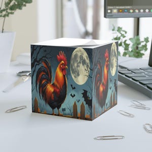May include: A square notepad featuring a vibrant rooster illustration against a spooky Halloween backdrop. The design includes a full moon, bats, and a wooden fence, creating a festive and eye-catching aesthetic. The rooster is depicted in shades of orange, red, and black.