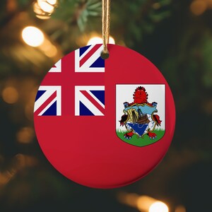 May include: A round Christmas ornament featuring the flag of Bermuda. The ornament is red with the Union Jack in the upper left corner and the Bermuda coat of arms on the right. A brown string is attached for hanging.