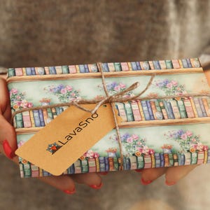 May include: A gift wrapped in paper with a bookshelf and floral design. The paper is light blue with brown shelves and colorful books. The gift is tied with twine and a brown tag that says "LavaSno".