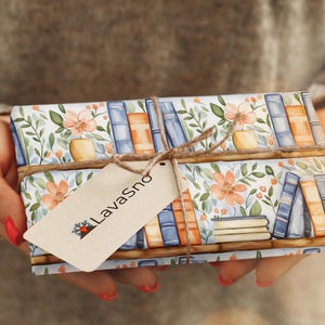 May include: A gift wrapped in white paper with a floral and book pattern. The paper is tied with twine and a brown tag with the text "LavaSno" printed on it.
