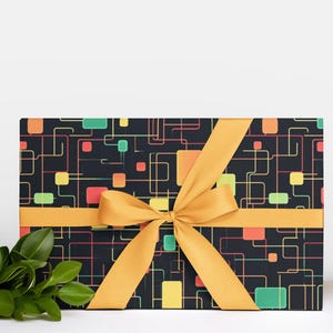 May include: A rectangular gift box wrapped in black paper with a colorful geometric pattern of squares and lines. The box is tied with a golden yellow ribbon and bow. Green leaves are visible on the left side.