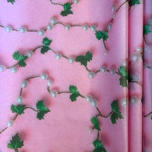 May include: Pink wrapping paper with a repeating pattern of white pearl strands and green leaf clusters. The paper has a soft, matte finish and a subtle texture, creating a delicate and elegant aesthetic.