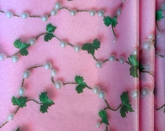 May include: Pink wrapping paper with a repeating pattern of white pearl strands and green leaf clusters. The paper has a soft, matte finish and a subtle texture, creating a delicate and elegant aesthetic.