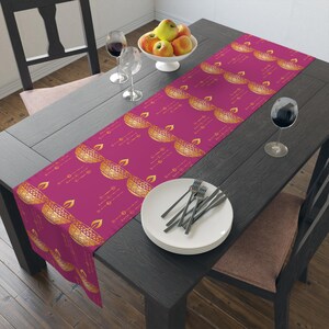 May include: A pink table runner with a gold diya pattern. The runner is on a dark wood table with a white plate and silverware.
