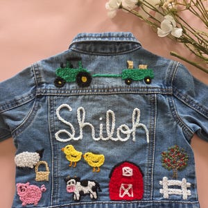May include: A blue denim jacket featuring colorful yarn embroidery on the back. The design includes a green tractor pulling a wagon, the name Shiloh in white yarn, a sheep, a pig, two yellow chicks, a cow, a red barn, a tree, and a white fence.