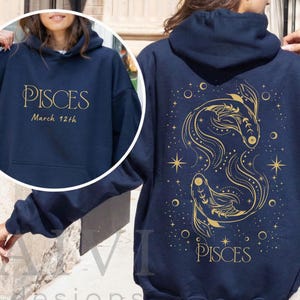 May include: Navy blue hoodie with gold text and graphics. The front features the word "PISCES" and "March 12th". The back displays a gold illustration of two fish, stars, and the word "PISCES".