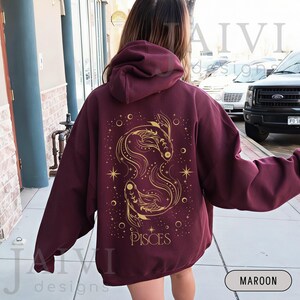 K&ouml;nnte beinhalten: Burgunderfarbener Kapuzenpullover mit goldenem Fische-Sternzeichen-Design auf dem R&uuml;cken. Das Design zeigt zwei Fische, Sterne und das Wort "PISCES". Der Hoodie hat eine Kapuze und lange &Auml;rmel.