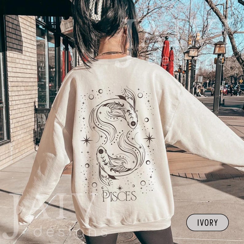 K&ouml;nnte beinhalten: Elfenbeinfarbenes Sweatshirt mit einem Fische-Sternzeichen-Design auf dem R&uuml;cken. Die Grafik zeigt zwei stilisierte Fische, Sterne und das Wort "PISCES" in einer dekorativen Schriftart. Das Sweatshirt hat einen Rundhalsausschnitt und lange &Auml;rmel.